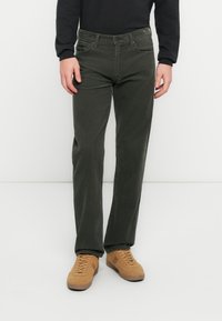 Jean vert foncé à jambes droites avec un tissu texturé, cinq poches et des ferrures en métal, associé à des baskets marron clair.