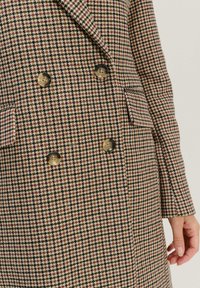 Dvostruki kaput u smešanoj, krem boji i maroon houndstooth uzorku, sa kontrastnim dugmadi i detaljem na džepu.