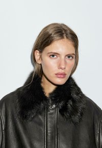 Giacca di pelle nera con colletto in pelliccia sintetica, con chiusura a bottoni e texture morbida. Il modello ha capelli lisci e un trucco neutro.