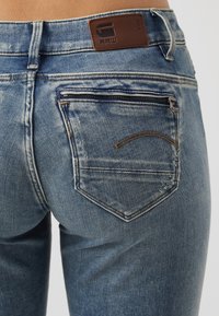 Close-up van de achterzak van blauwe spijkerbroek met rits en bruine leren merkpatch op de tailleband.