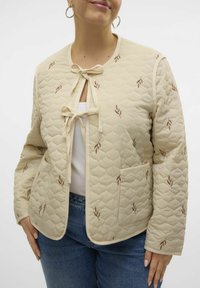 Beige gewatteerde jas met een striksluiting, voorzien van bruine bladeren borduurwerk. Het heeft een rond halslijn en een zak aan de linkerkant.