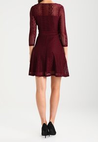 Bordeaux kanten jurk met driekwart mouwen, strakke taille, uitlopende rok en ingewikkelde patronen; gecombineerd met zwarte high heels.