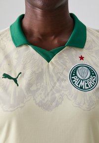 Dzeltena sporta krekls ar zaļu apkakli. Satur Puma logo un apļa Palmeiras emblēmu ar sarkanu zvaigzni. Tekstūras audums ar ziedu rakstu.