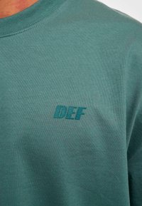 Teal bomull T-skjorte med rund hals og en teksturert "DEF" logo i matchende teal-farge på brystet. Myk og avslappet design.