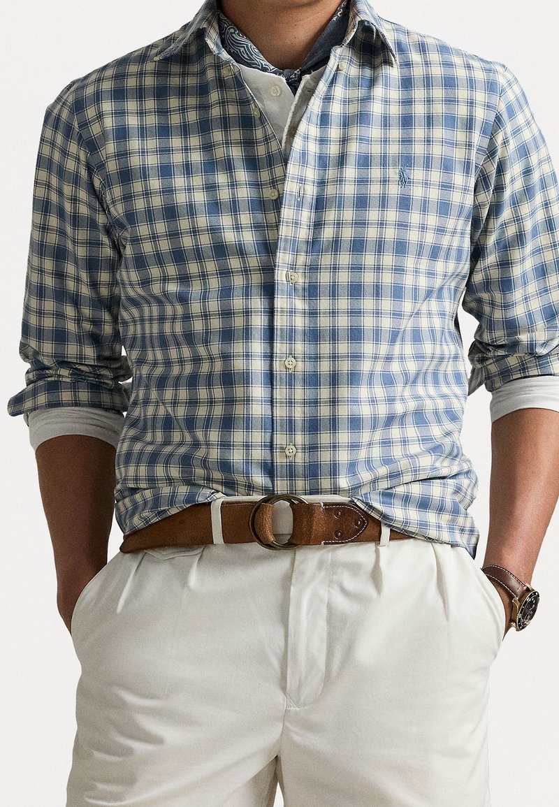 Sininen ja valkoinen ruudullinen nappipaita, pitkät hihat käärittyinä, yhdistettynä kermaisiin housuihin ja ruskeaan nahkavyöhön. Huomiota herättävä rannekello.