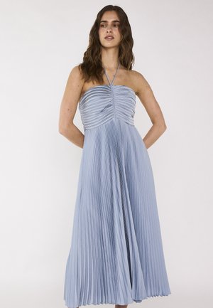 Femme aux cheveux bruns ondulés portant une robe plissée bleu clair sans bretelles avec un col halter, se tenant les mains derrière le dos.