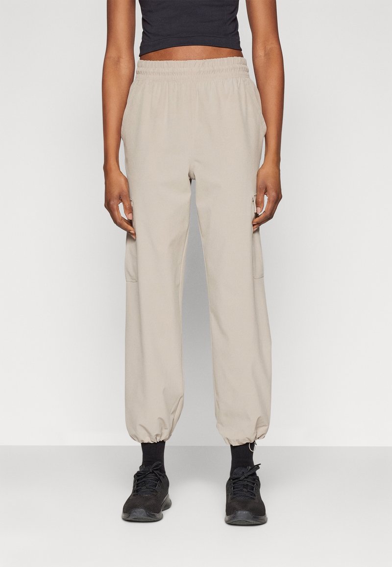 Pantaloni cargo beige con vita ed orli elastici, dotati di tasche laterali e una texture liscia, indossati con scarpe nere e calze.