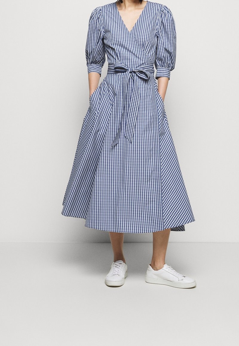 Blå och vit rutig klänning med omslagsdesign, puffärmar och ett knytbälte. Har en full kjol och sidofickor. I kombination med vita sneakers.