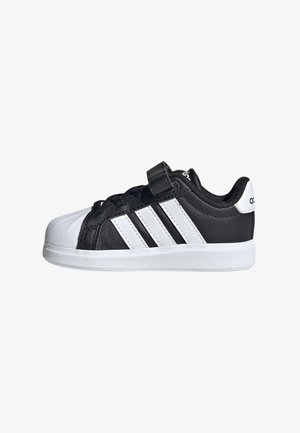 Zapatilla Adidas en blanco y negro con tres franjas blancas, correa de velcro, cordones negros, suela blanca y punta redondeada.