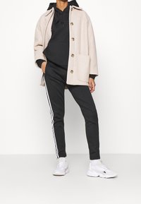 Beige mantel van een wolmix met grote knopen over een zwarte hoodie. Zwarte joggers met witte zijstrepen en witte sneakers.