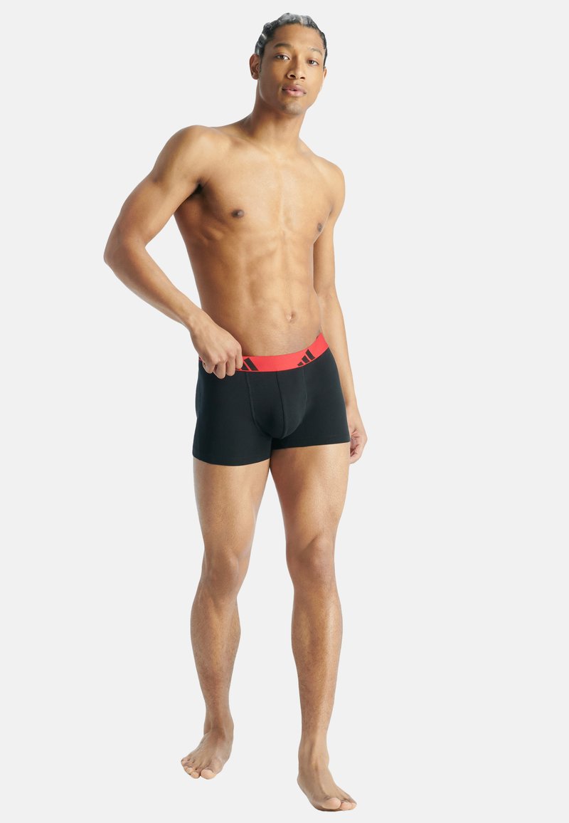 Černé boxerky s červeným elastickým pasem, na kterém je logo. Materiál vypadá hladce a přiléhá k tělu, zdůrazňuje jeho tvar.