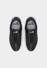 Ζευγάρι μαύρα αθλητικά παπούτσια Love Moschino με χρυσές τρύπες για κορδόνια, μαύρα κορδόνια και λευκές σόλες, εμφανιζόμενα από πάνω σε απλό φόντο.