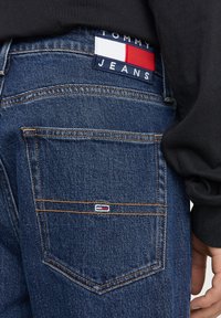 Jeans de mezclilla azules con costuras naranjas, que presentan una etiqueta en la parte trasera que dice "TOMMY JEANS" en azul marino, blanco y rojo, y un pequeño logotipo en el bolsillo.