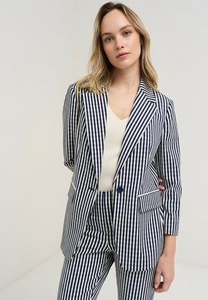 Kvinde med skulderlangt blondt hår iført en mørkeblå og hvide lodrette stribede blazer og matchende bukser, kombineret med en hvid top.