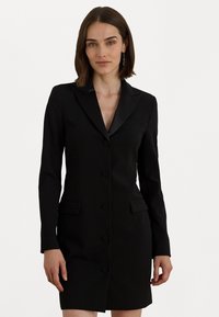 Žena oblečená v priliehavých čiernych saku šatách s dlhými rukávmi a piatimi zakrytými gombíkmi, stojaca na pozadí jednoduchého bieleho pozadia.