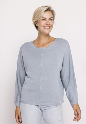 Smiling woman with short blonde hair wearing a light blue knit sweater and light washed jeans, standing against a plain background.