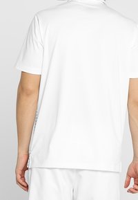 Personne portant un polo blanc à manches courtes et un short blanc, vue de dos sur un fond uni.