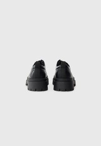 Zapatos de cuero negro con suela gruesa, que presentan una textura suave y una lengüeta en la parte trasera para facilitar su calzado. Diseño elegante y minimalista.