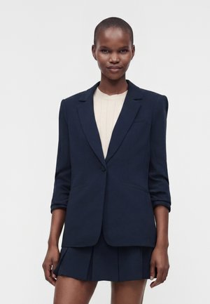 Vrouw met kort haar draagt een marineblazer over een crème top en bijpassende marineblauwe geplooide rok tegen een effen grijze achtergrond.