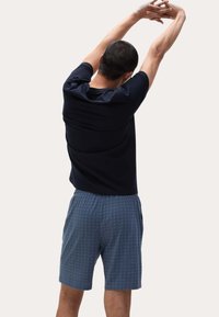 Hombre estirando los brazos por encima de la cabeza, vistiendo una camiseta negra y pantalones cortos azules con estampado, de pie frente a un fondo claro y liso.