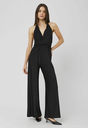 Femme portant une combinaison noire sans manches à col halter avec jambes larges et des talons noirs pointus, posant avec une main sur la hanche.