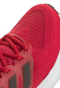 Zapatilla deportiva roja con parte superior de punto texturizado, sistema de cordones prominente y suela de goma blanca con patrón de tracción. Destacan los acentos en negro.