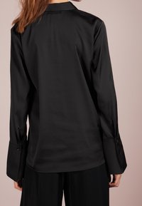 Camicia nera in satin con maniche lunghe, colletto alto e polsini abbottonati; texture liscia e design minimalista.