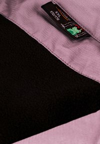 Lila und schwarzes Fleece-Innenfutter mit glatter Textur, versehen mit einem Etikett mit einem grünen Frosch-Logo und Größeninformationen.