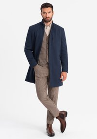 Cappotto blu navy con colletto alto, tasca con zip, indossato su una camicia beige e un gilet a scacchi, abbinato a stivali marroni e pantaloni con motivo.