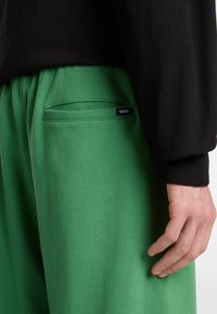 Groene katoenen trainingsbroek met zijzak en Vans-logo, gecombineerd met een zwart longsleeve shirt. Close-up die texturen en stiksels toont.