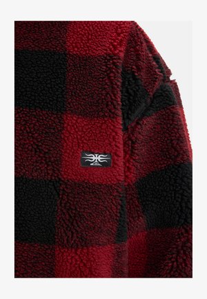 Rote und schwarze Karomuster-Fleecejacke mit einer strukturierten Oberfläche und einem Logo-Aufnäher auf der Vorderseite. Weiches, gemütliches Material, das warm hält.