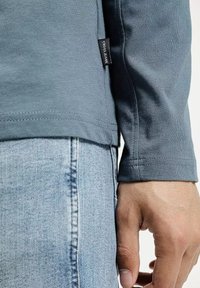 Zbliżenie na osobę w niebieskiej bluzie z długim rękawem z metką "Cross Jeans" oraz jasnoniebieskich dżinsach, ręka swobodnie leży na boku.