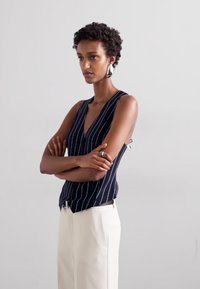 Mornarsko modra in bela črtasta brezrokavnica z globokim V-izrezom in stranskimi razpoki, v kombinaciji z visokimi belimi hlačami. Preprost dizajn, gladka tekstura.