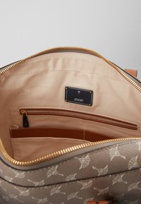 Beige-fôret håndtaske med lynlås lomme, åben rum, guldlås og sort label med teksten "JOOP!" på det mønstrede ydre.