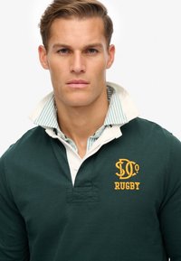 Mørkegrøn rugbytrøje med hvid krave og stribet indvendig foring. Har gule broderede "RUGBY"-tekst og quiltet placket-detalje.