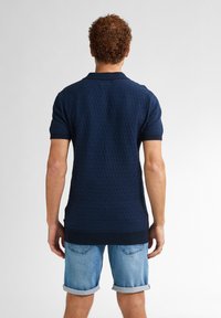 Námořnická modrá polo košile s texturovaným vlnkovým vzorem, žebrovaným límcem a manžetami, v kombinaci se světle modrými ohrnutými džínovými šortkami.