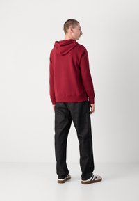 Piros kapucnis hoodie, laza fazonnal és állítható kapucnival, sötétszürke laza fazonú nadrággal, valamint fekete részletekkel ellátott fehér sportcipőkkel párosítva.