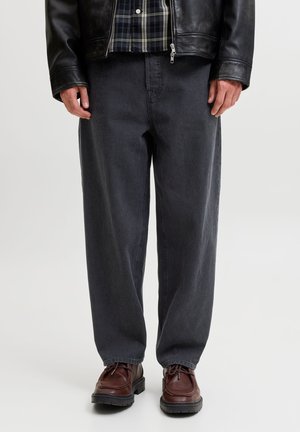 Homme portant un jean ample gris foncé, des chaussures en cuir marron, une veste en cuir noire et une chemise à carreaux noir et blanc, debout sur un sol blanc.