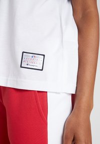 Camiseta blanca de manga corta con una etiqueta de MLB en azul y rojo que muestra "Mercancía Genuina de Champion", combinada con pantalones rojos.