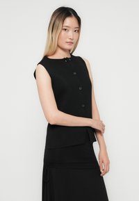 Kvinna med långt rakt hår som bär en ärmlös svart knappad topp och svart kjol, stående mot en enhetlig ljus bakgrund.