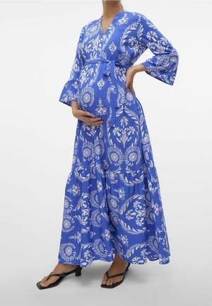 Kobieta w ciąży ubrana w długą sukienkę kopertową w niebiesko-białe kwiaty, trzymająca jedną ręką brzuch, a drugą na biodrze, stojąca w czarnych sandałach na obcasie.