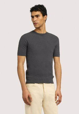 Uomo con capelli ricci scuri che indossa una maglietta a maniche corte grigio scuro con texture e pantaloni beige chiaro, in piedi contro uno sfondo chiaro e semplice.