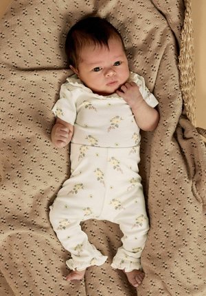 Nouveau-né aux cheveux foncés allongé sur une couverture beige texturée, portant une tenue blanche à fleurs avec un pantalon à volants.