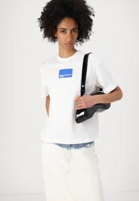Cămașă albă cu logo grafic albastru, asortată cu o fustă din denim deschis. Modelul ține o geantă mică, neagră, lucioasă, cu un design curbat.