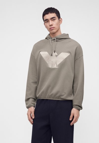 Jeune homme portant un sweat à capuche taupe avec un logo d'aigle et les lettres GA sur le devant ainsi qu'un pantalon sombre, debout devant un fond uni.