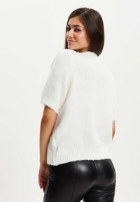 White short-sleeve sweater with a textured knit, featuring a ribbed hem. Paired with black fitted pants, showcasing a sleek silhouette.
