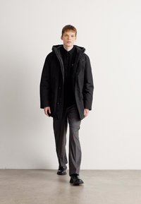 Čierny kapucňový parkas so zipsom, nosený cez čiernu košelu a šedé vzorované nohavice. Model stojí na jednoduchom pozadí.
