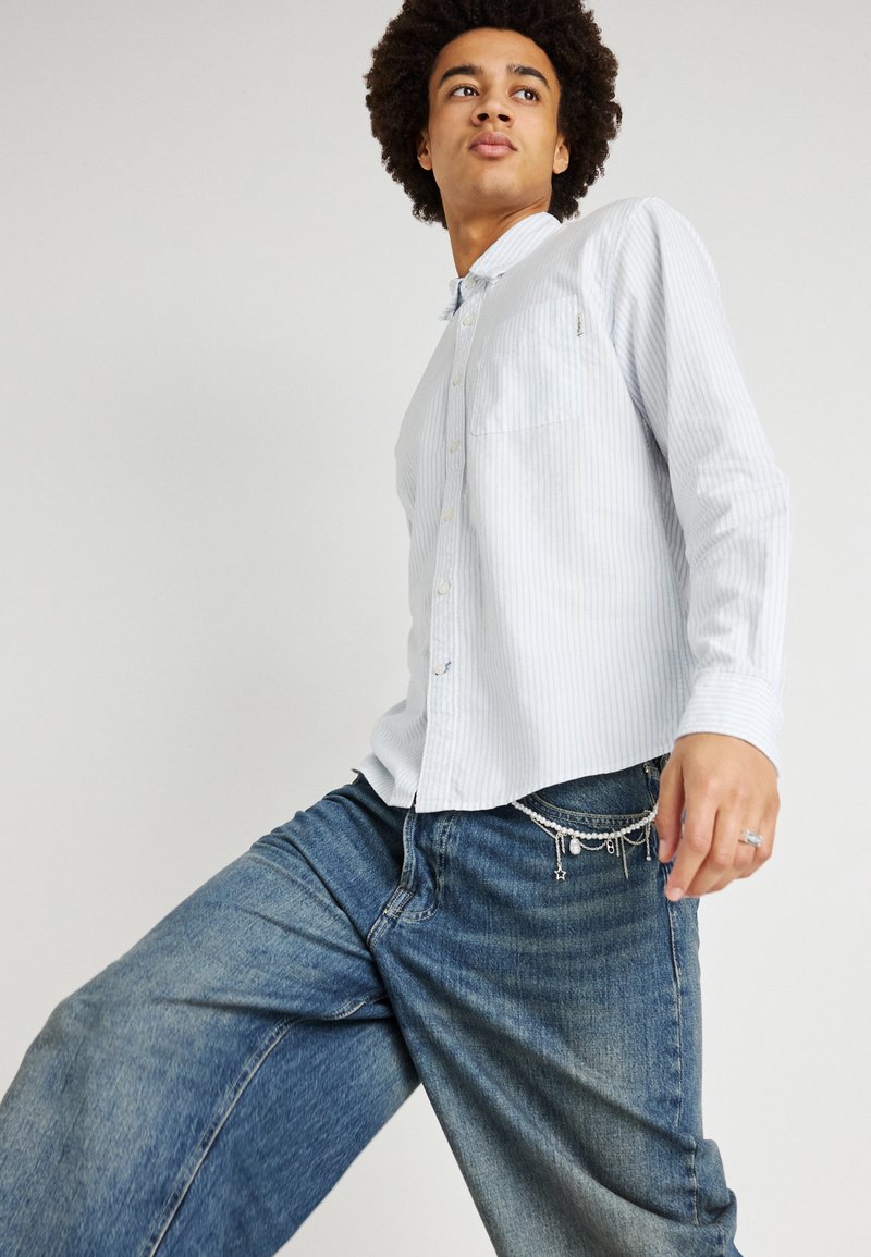 Ανοιχτό μπλε πουκάμισο με λεπτές λευκές ρίγες, μακριά μανίκια και τσέπη στο στήθος, σε συνδυασμό με φαρδιά μπλε τζιν με αλυσίδες.