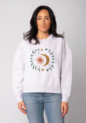 Frau mit dunklem Haar, die einen hellgrauen Pullover mit einem Sonnen- und Mondgrafik trägt, umgeben von grünen Blättern, kombiniert mit blauen Jeans.