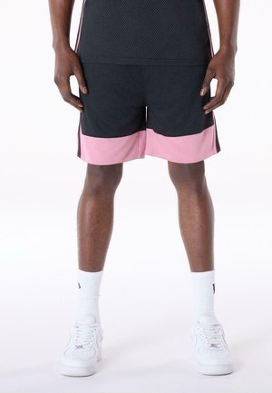 Person trägt schwarze und pinke Sportshorts, weiße Crew-Socken und weiße Sneakers und steht auf einer weißen Fläche.